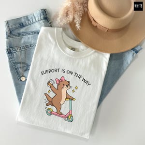 May include: A white t-shirt featuring a graphic of a brown otter wearing a pink bow, riding a pink and blue scooter while holding a coffee cup. The text "SUPPORT IS ON THE WAY" is printed above the otter. The shirt is paired with blue jeans.