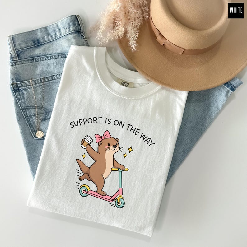 May include: A white t-shirt featuring a graphic of a brown otter wearing a pink bow, riding a pink and blue scooter while holding a coffee cup. The text "SUPPORT IS ON THE WAY" is printed above the otter. The shirt is paired with blue jeans.