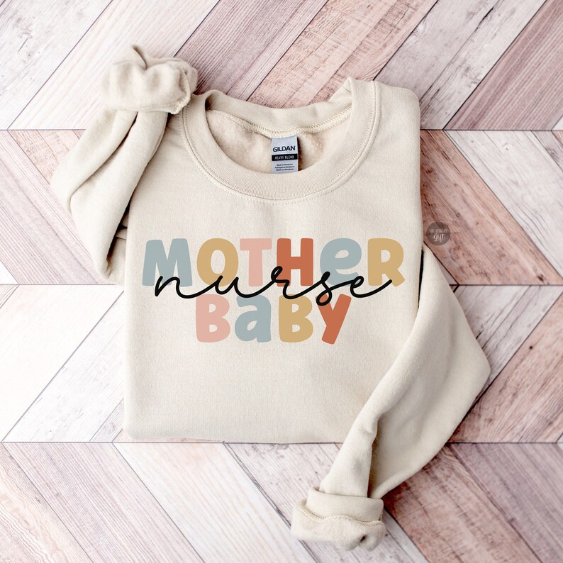 Pode incluir: Uma camisa de moletom bege com o texto "MOTHER nurse BABY" em letras de bloco coloridas. As letras s&atilde;o em tons de rosa, azul, amarelo e laranja.
