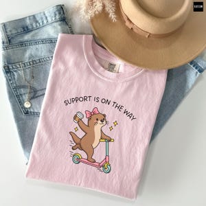 May include: A light pink t-shirt featuring a graphic of a brown otter wearing a pink bow, riding a pink and teal scooter while holding a coffee cup. The text "SUPPORT IS ON THE WAY" is printed above the otter. The shirt is paired with blue jeans.