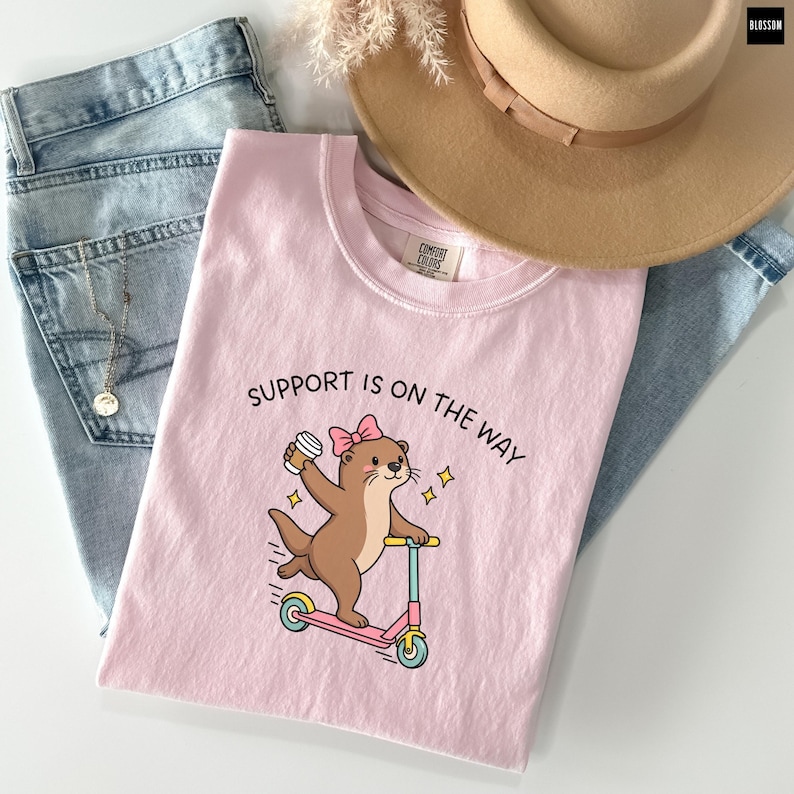 May include: A light pink t-shirt featuring a graphic of a brown otter wearing a pink bow, riding a pink and teal scooter while holding a coffee cup. The text "SUPPORT IS ON THE WAY" is printed above the otter. The shirt is paired with blue jeans.