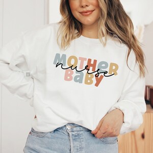 Pode incluir: Camisola branca com o texto "MOTHER nurse BABY" em letras coloridas. As letras est&atilde;o em um esquema de cores arco-&iacute;ris.