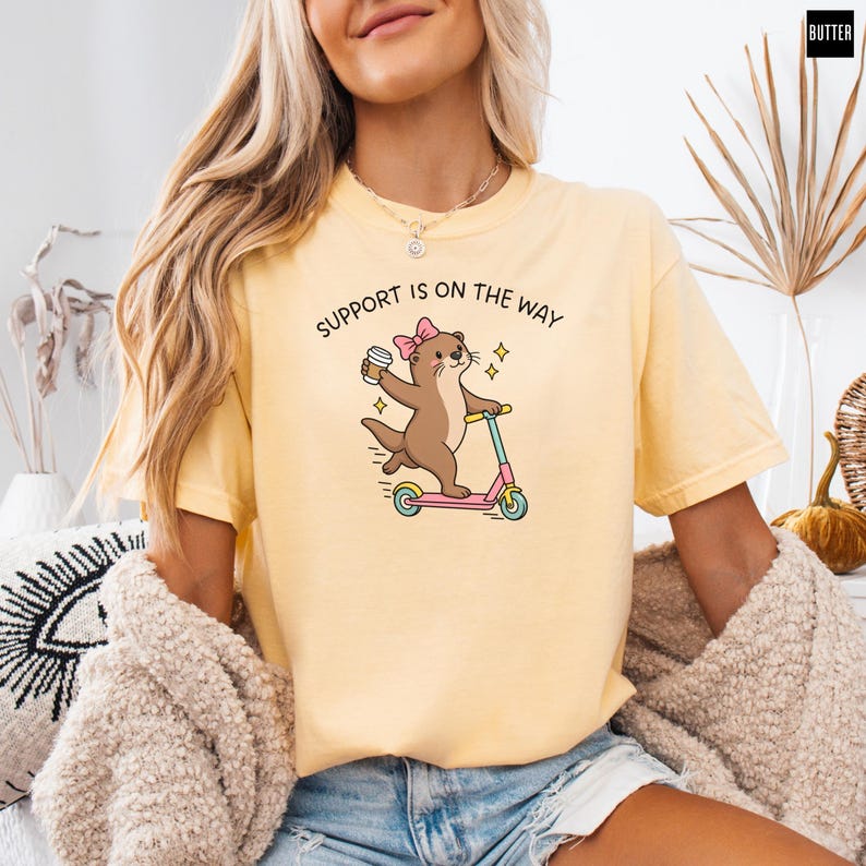 May include: A pale yellow short-sleeve t-shirt featuring a brown otter wearing a pink bow, riding a blue and pink scooter while holding a coffee cup. The text above the otter reads SUPPORT IS ON THE WAY in black capital letters.