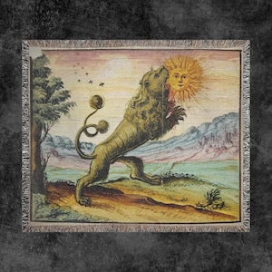 Puede incluir: Una manta tejida con una ilustración vintage de un león mordiendo un sol con una cara. La obra de arte está ambientada en un paisaje con un árbol, montañas y un castillo. La manta tiene un borde con flecos.