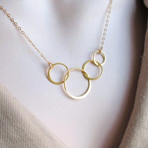 Puede incluir: Un delicado collar dorado con cinco círculos interconectados de varios tamaños. Los círculos están suspendidos de una fina cadena, creando un diseño minimalista y elegante. El collar se muestra sobre un fondo neutro.