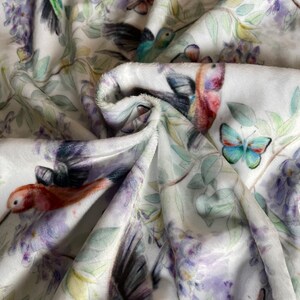 May include: White fabric with a repeating pattern of hummingbirds, butterflies, and purple flowers. The fabric is soft and plush.