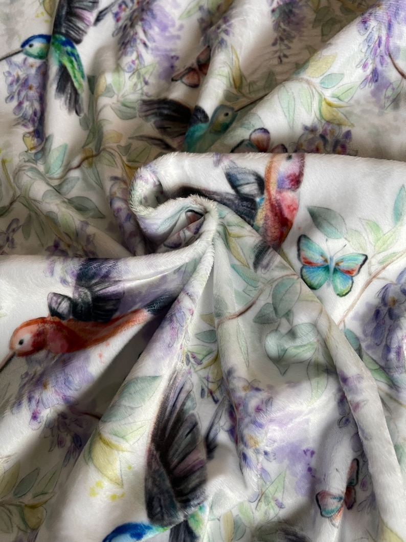 May include: White fabric with a repeating pattern of hummingbirds, butterflies, and purple flowers. The fabric is soft and plush.