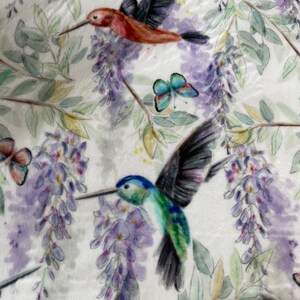 May include: A white fabric with a repeating pattern of hummingbirds, butterflies, and wisteria flowers. The hummingbirds are depicted in shades of red, green, and blue, while the butterflies are blue and purple. The wisteria flowers are a light purple colour.