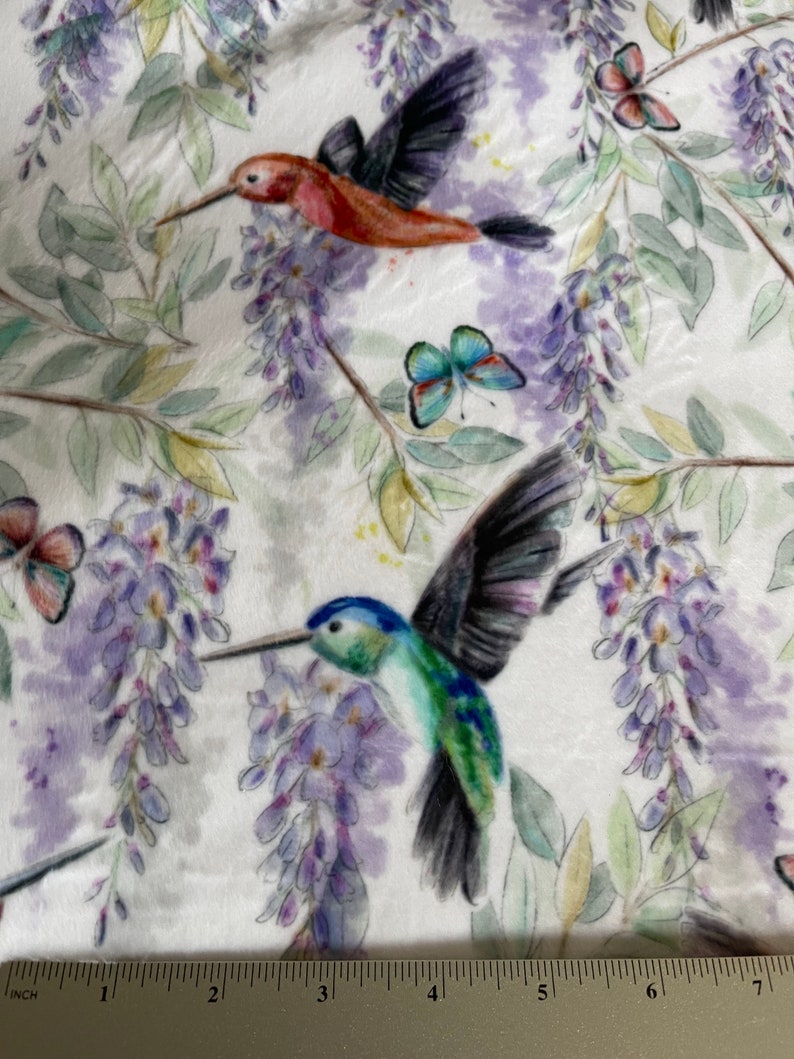 May include: A white fabric with a repeating pattern of hummingbirds, butterflies, and wisteria flowers. The hummingbirds are depicted in shades of red, green, and blue, while the butterflies are blue and purple. The wisteria flowers are a light purple colour.