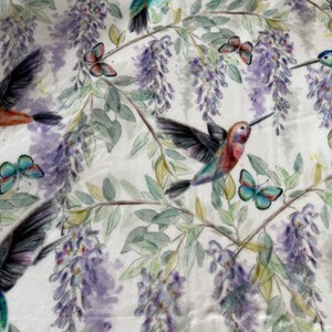 May include: A white fabric with a repeating pattern of hummingbirds, butterflies, and wisteria flowers. The hummingbirds are in shades of blue, green, and red. The butterflies are in shades of blue, green, and pink. The wisteria flowers are in shades of purple and white.
