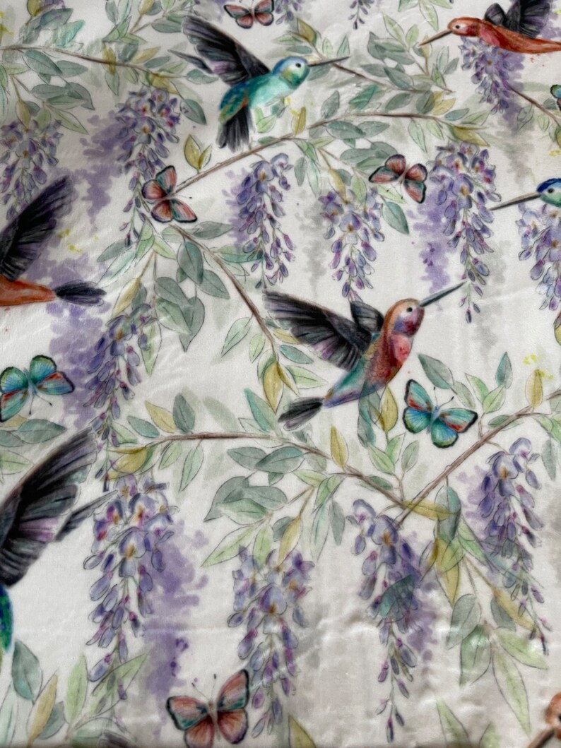 May include: A white fabric with a repeating pattern of hummingbirds, butterflies, and wisteria flowers. The hummingbirds are in shades of blue, green, and red. The butterflies are in shades of blue, green, and pink. The wisteria flowers are in shades of purple and white.