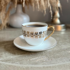 May include: A white teacup and saucer set with gold trim and a gold geometric pattern. The cup is filled with dark liquid, likely coffee or tea. The handle is gold. The set is on a light-colored wooden surface.