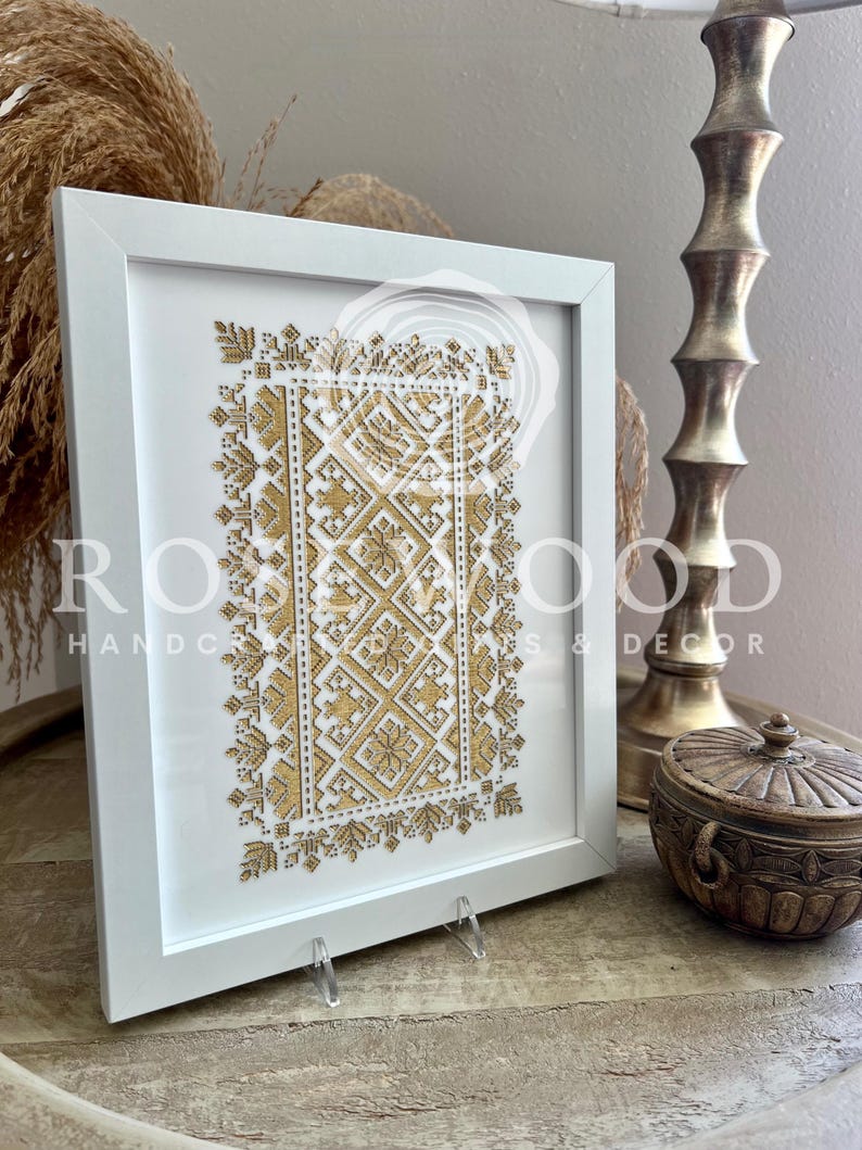 K&ouml;nnte beinhalten: Ein wei&szlig; gerahmtes Bild mit einem goldenen Stickmuster. Das Muster ist ein geometrisches Muster mit einem zentralen Quadrat und vier kleineren Quadraten darum herum. Das Bild steht auf einem Holztisch.