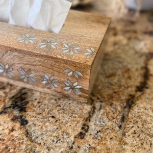 May include: A rectangular wooden tissue box with a light brown finish. The box is decorated with inlaid floral designs in a lighter color. White tissues are visible at the top. The box sits on a speckled, brown surface.