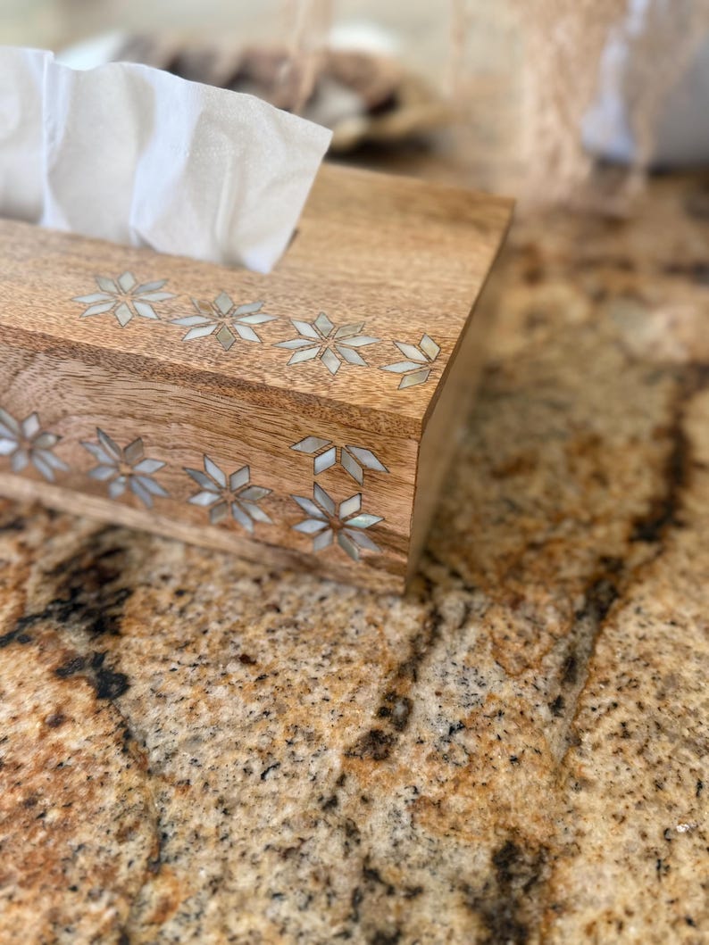 May include: A rectangular wooden tissue box with a light brown finish. The box is decorated with inlaid floral designs in a lighter color. White tissues are visible at the top. The box sits on a speckled, brown surface.