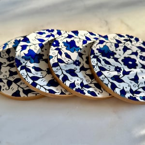May include: Set of four round coasters with a white background and a blue floral design. The coasters have a wooden rim and feature a repeating pattern of blue flowers and leaves. These coasters are perfect for protecting surfaces from drink rings.