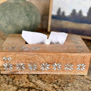 May include: Wooden tissue box with inlaid starburst designs. The box is rectangular with a natural wood finish and a slot for tissue dispensing. Decorative accents include mother-of-pearl starbursts along the sides and top.