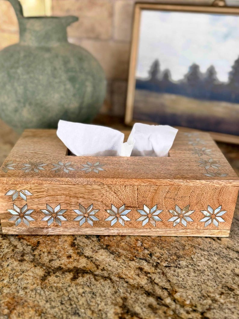 May include: Wooden tissue box with inlaid starburst designs. The box is rectangular with a natural wood finish and a slot for tissue dispensing. Decorative accents include mother-of-pearl starbursts along the sides and top.