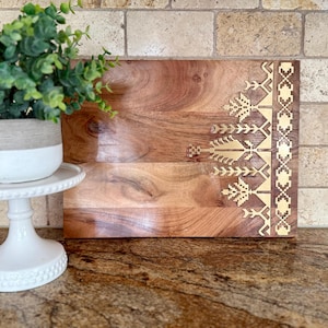 May include: A wooden cutting board with a gold geometric design on the edge. The board is made of natural wood and has a warm brown color.
