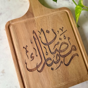 May include: Wooden cutting board with a handle and a rectangular cutting surface. The board has a carved Arabic inscription in a dark brown color. The board is made of light-colored wood.