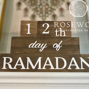 May include: A wooden block calendar with the text "12th day of RAMADAN" in white lettering. The blocks are arranged in a tiered design, with the brand name "ROSEWOOD HANDCRAFTED DESIGNS" visible in the background.