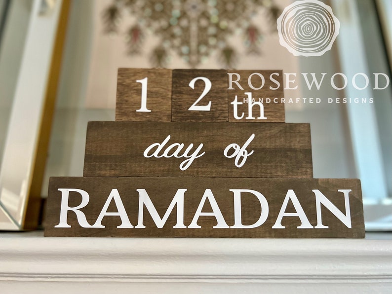 May include: A wooden block calendar with the text "12th day of RAMADAN" in white lettering. The blocks are arranged in a tiered design, with the brand name "ROSEWOOD HANDCRAFTED DESIGNS" visible in the background.
