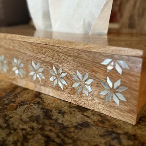 May include: Wooden tissue box with a glossy finish. The box is decorated with inlaid mother-of-pearl starburst designs along the front. White tissues are visible, pulled from the top opening. The box sits on a speckled brown surface.