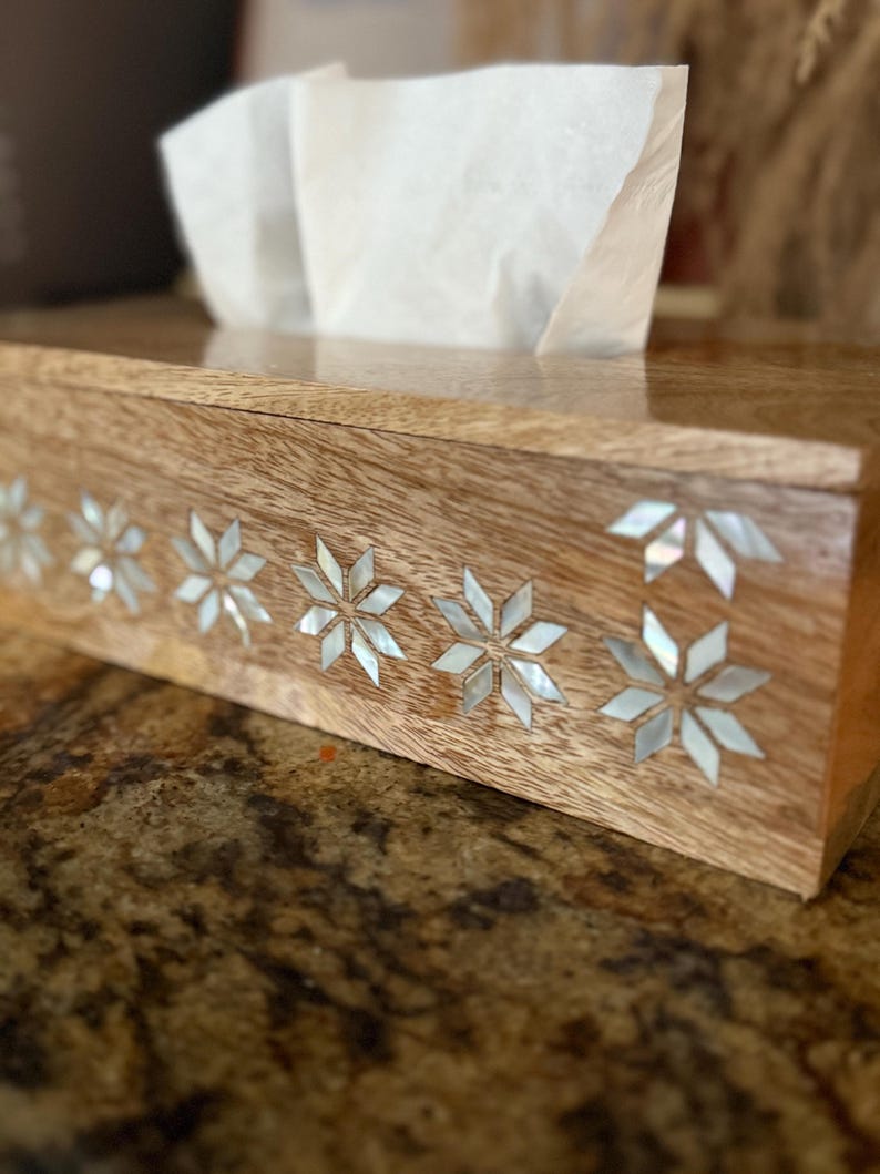 May include: Wooden tissue box with a glossy finish. The box is decorated with inlaid mother-of-pearl starburst designs along the front. White tissues are visible, pulled from the top opening. The box sits on a speckled brown surface.