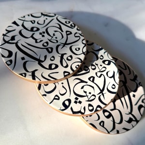 Puede incluir: Tres posavasos redondos con fondo blanco y diseño de caligrafía árabe negra. Los posavasos tienen un borde de madera marrón claro. Los posavasos están apilados ligeramente superpuestos.