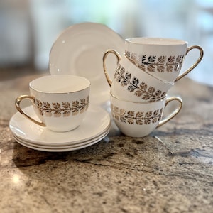 May include: A set of white teacups and saucers with gold trim and a gold floral pattern. The teacups are stacked, and a plate is in the background. The teacups have gold handles.