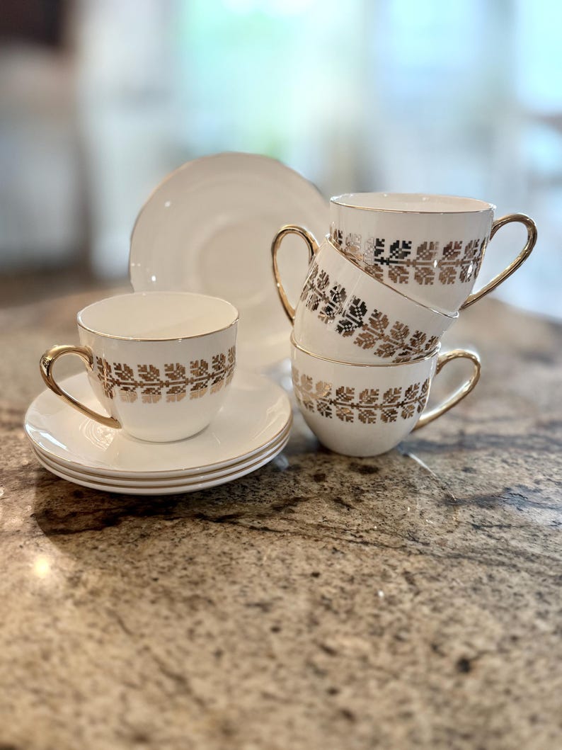 May include: A set of white teacups and saucers with gold trim and a gold floral pattern. The teacups are stacked, and a plate is in the background. The teacups have gold handles.