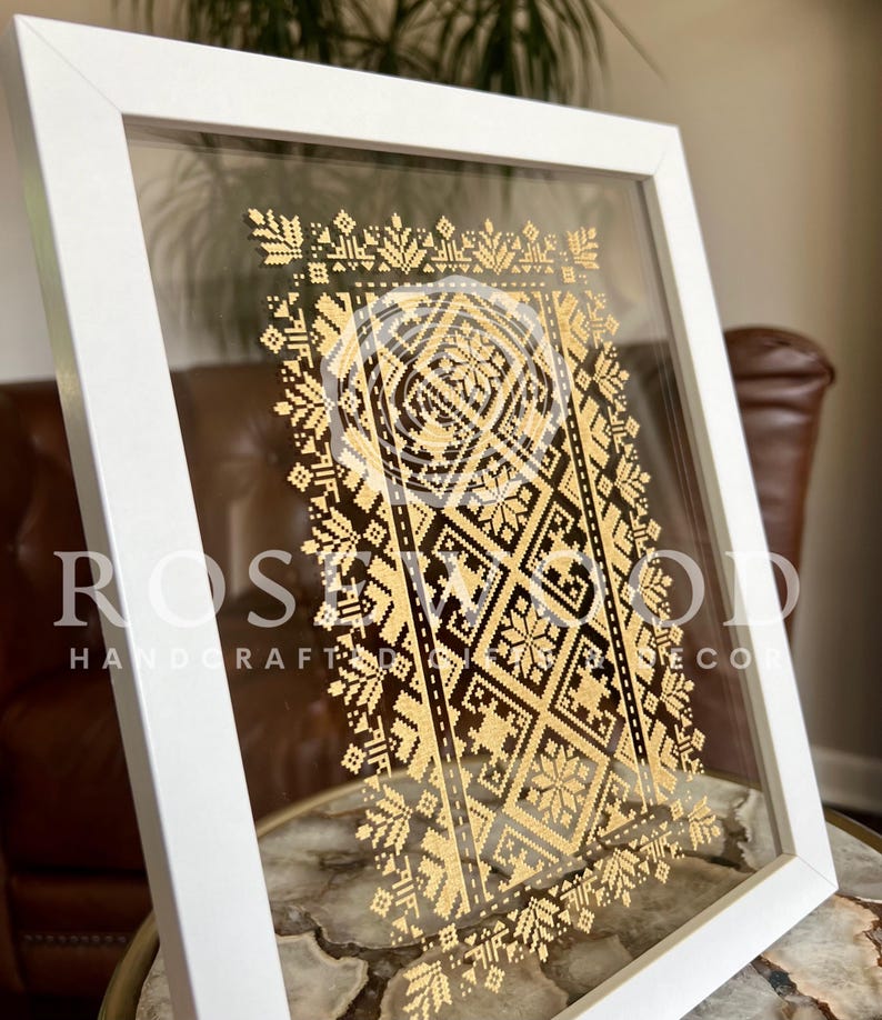 K&ouml;nnte beinhalten: Ein wei&szlig; gerahmtes Bild mit einem goldenen geometrischen Muster im Inneren. Das Muster ist ein traditionelles ukrainisches Design. Der Rahmen ist aus Holz und hat eine Glasfront.