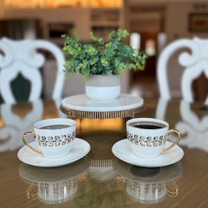 May include: Two white teacups and saucers with gold trim and floral designs, filled with dark liquid, sit on a reflective surface. A small plant in a white and gray pot rests on a decorative stand in the background. The scene suggests a tea party setting.