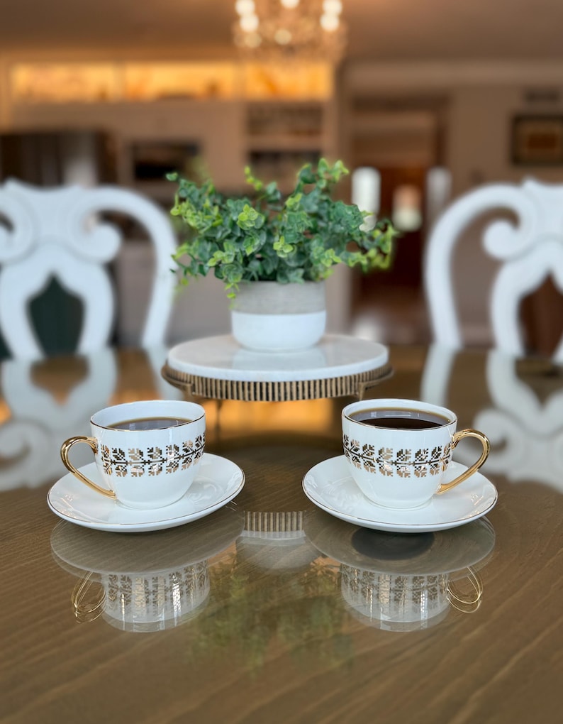 May include: Two white teacups and saucers with gold trim and floral designs, filled with dark liquid, sit on a reflective surface. A small plant in a white and gray pot rests on a decorative stand in the background. The scene suggests a tea party setting.