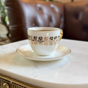 May include: A white teacup and saucer set with gold trim and a decorative gold floral pattern. The teacup is filled with dark liquid and sits on a white saucer. The cup has a gold handle. The set is on a white marble table.