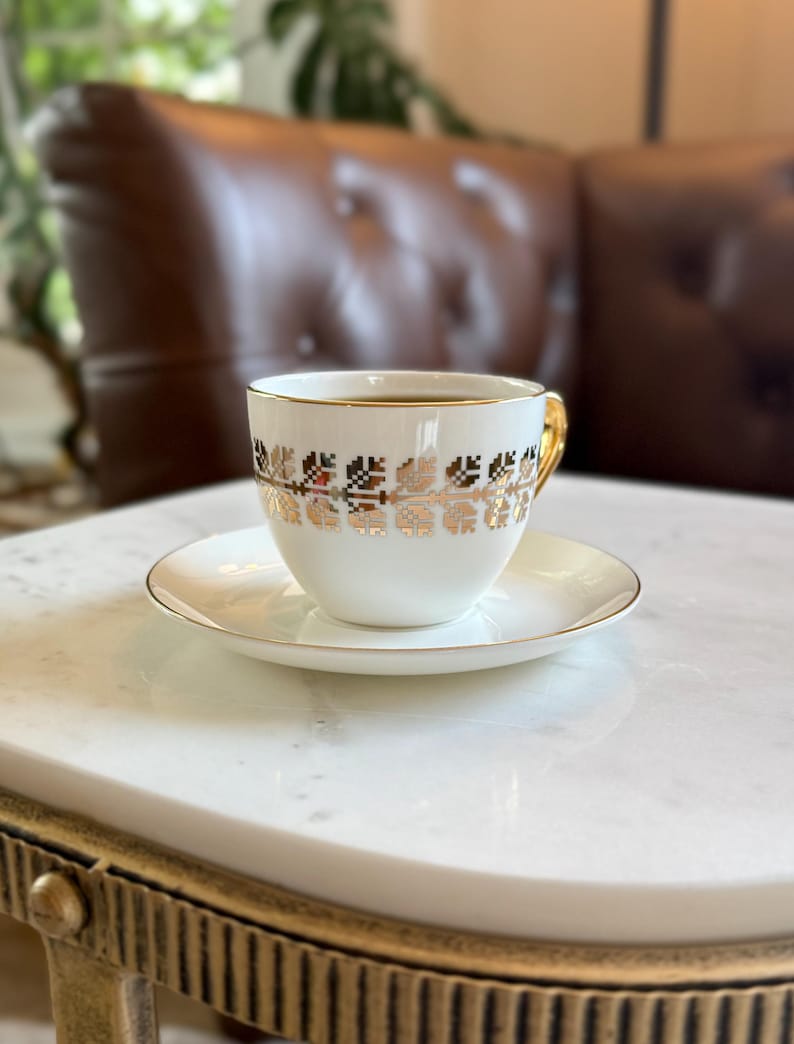 May include: A white teacup and saucer set with gold trim and a decorative gold floral pattern. The teacup is filled with dark liquid and sits on a white saucer. The cup has a gold handle. The set is on a white marble table.