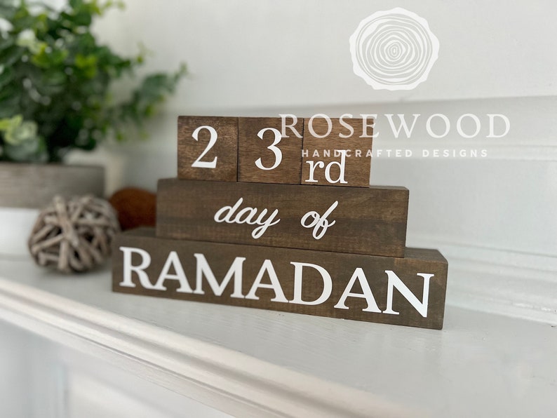May include: A wooden block calendar with the text "23rd day of RAMADAN" in white lettering. The blocks are stacked, with the numbers and text clearly visible against the dark brown wood. The background includes greenery and decorative items.