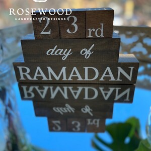 May include: Wooden block calendar displaying "23rd day of Ramadan" in white lettering. The blocks are arranged in a tiered design, with the text clearly visible. The Rosewood Handcrafted Designs logo is at the top.