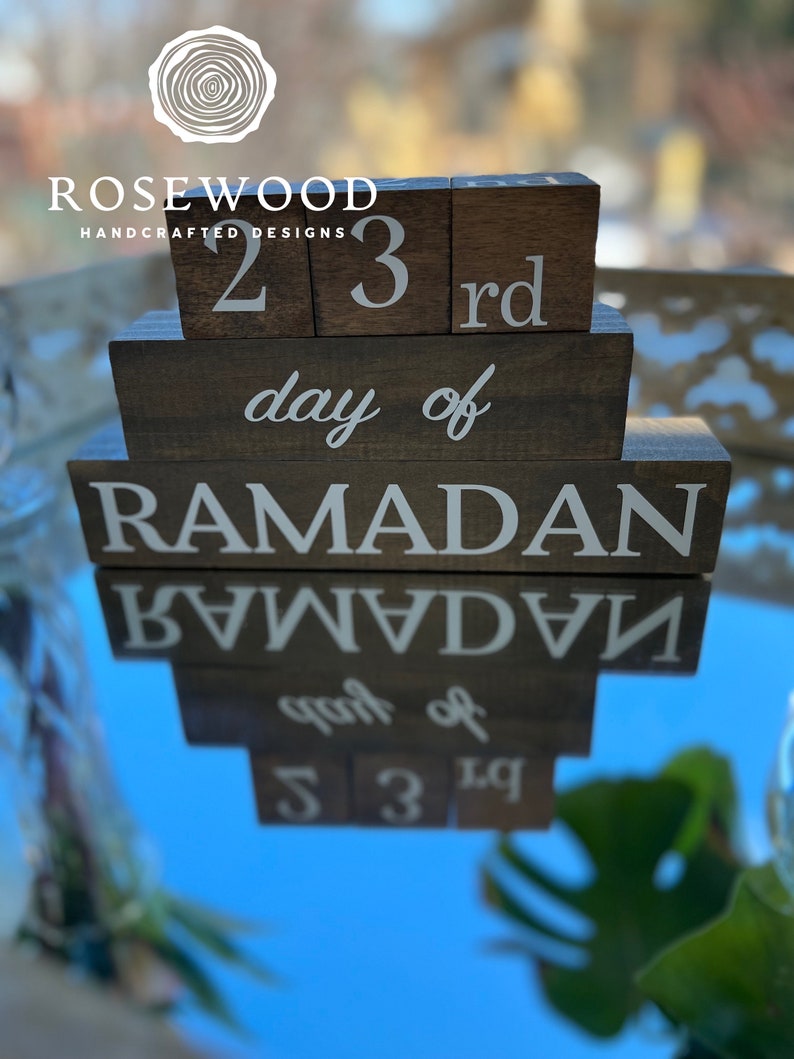 May include: Wooden block calendar displaying "23rd day of Ramadan" in white lettering. The blocks are arranged in a tiered design, with the text clearly visible. The Rosewood Handcrafted Designs logo is at the top.