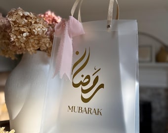 May include: A white plastic gift bag with a gold handle and a gold ribbon. The bag has a gold inscription that says "Ramadan Mubarak".