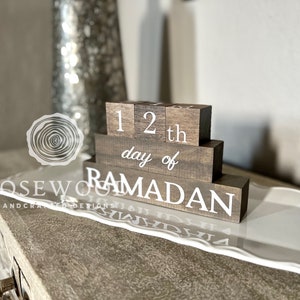 May include: A wooden block calendar displays "12th day of Ramadan" in white lettering. The blocks are stacked on a white decorative tray, with a blurred background featuring a vase and a light-colored surface.