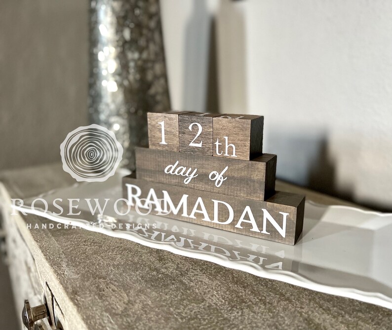May include: A wooden block calendar displays "12th day of Ramadan" in white lettering. The blocks are stacked on a white decorative tray, with a blurred background featuring a vase and a light-colored surface.