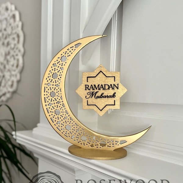May include: A gold wooden crescent moon with intricate cutouts and a star-shaped sign that reads "RAMADAN Mubarak" hanging from it. The crescent moon is on a white surface.