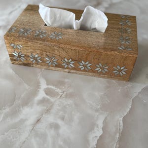 May include: A rectangular wooden tissue box with a light brown finish. The box is decorated with inlaid floral patterns along the edges. White tissues are visible, pulled from the top opening. The box sits on a light-colored, marble-like surface.