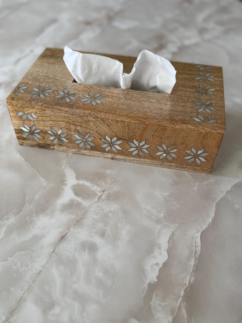 May include: A rectangular wooden tissue box with a light brown finish. The box is decorated with inlaid floral patterns along the edges. White tissues are visible, pulled from the top opening. The box sits on a light-colored, marble-like surface.