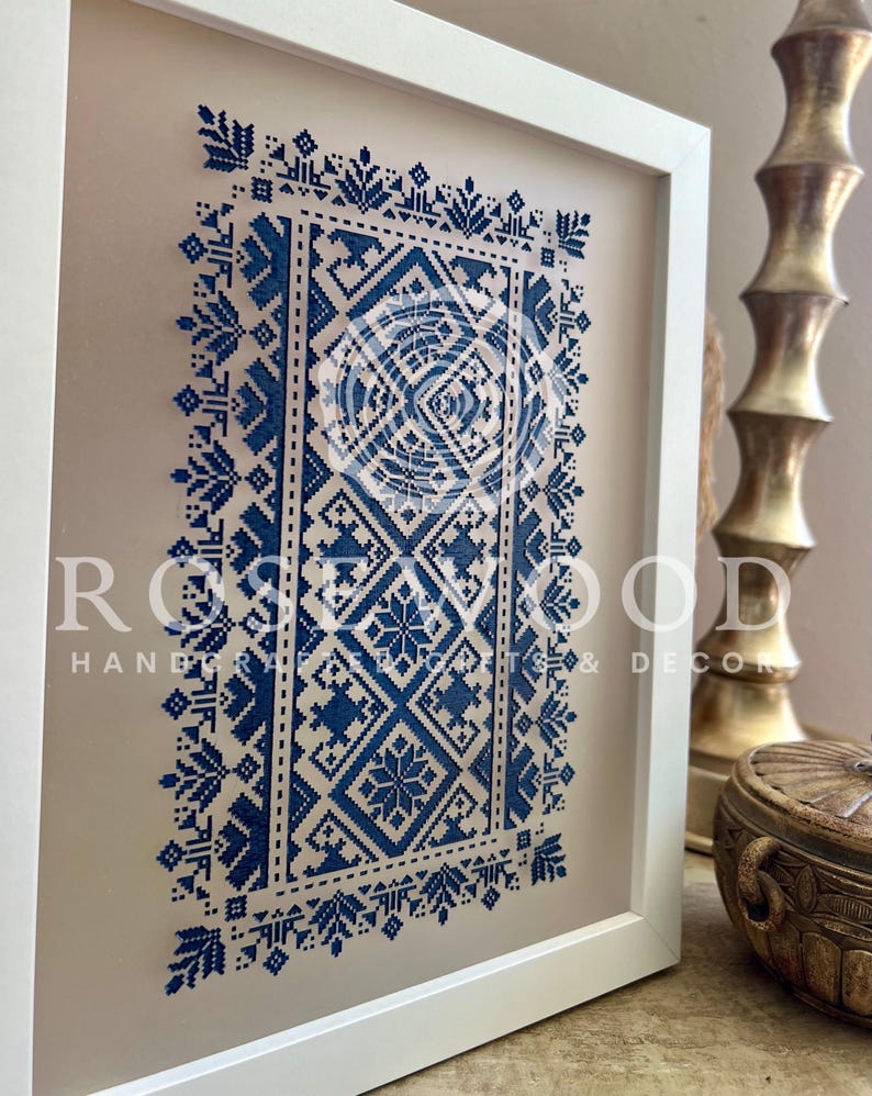 K&ouml;nnte beinhalten: Eine gerahmte blaue und wei&szlig;e geometrische Muster-Kreuzstich-Stickerei. Die Stickerei befindet sich in einem wei&szlig;en Rahmen mit einer beigefarbenen Passepartout. Der Text "ROSEWOOD HANDCRAFTED & DECOR" ist in der unteren rechten Ecke des Bildes sichtbar.