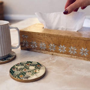 May include: A wooden tissue box with inlaid star designs, a white mug, and a coaster with a green and gold pattern sit on a table. A hand reaches for a tissue. The items are on a light-colored surface.