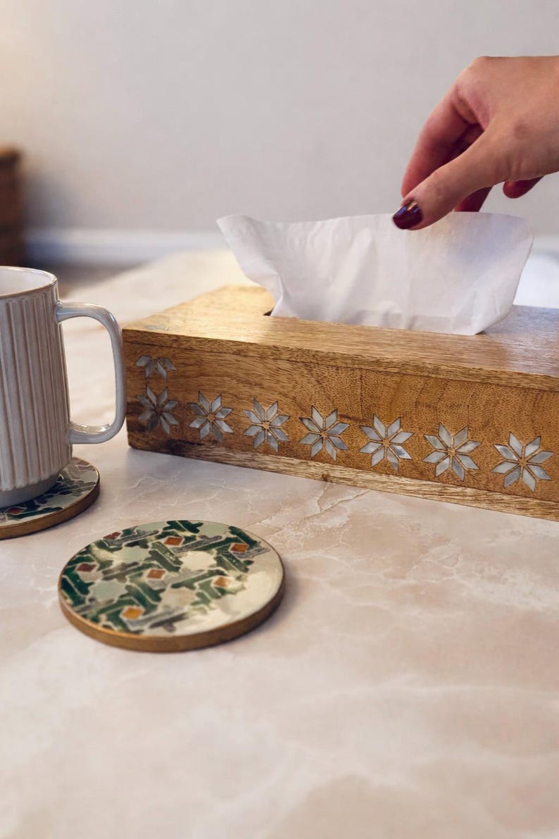 May include: A wooden tissue box with inlaid star designs, a white mug, and a coaster with a green and gold pattern sit on a table. A hand reaches for a tissue. The items are on a light-colored surface.