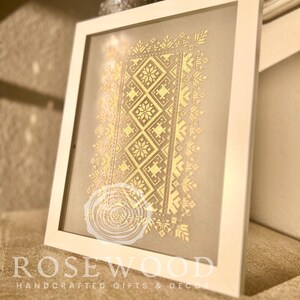 K&ouml;nnte beinhalten: Ein gerahmtes Stickbild mit einem goldenen geometrischen Muster auf beigefarbenem Hintergrund. Der Rahmen ist wei&szlig; und der Text "ROSEWOOD HANDCRAFTED GIFTS & DECOR" befindet sich unten.