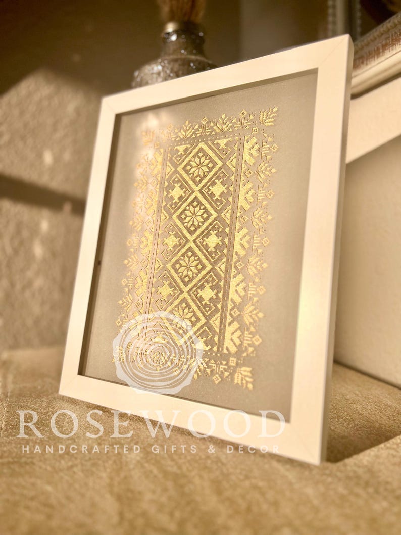 K&ouml;nnte beinhalten: Ein gerahmtes Stickbild mit einem goldenen geometrischen Muster auf beigefarbenem Hintergrund. Der Rahmen ist wei&szlig; und der Text "ROSEWOOD HANDCRAFTED GIFTS & DECOR" befindet sich unten.